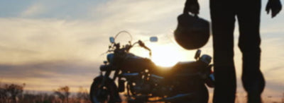 A motorcycle rider walking away from their parked bike with the sun setting behind it | Eagle Leather