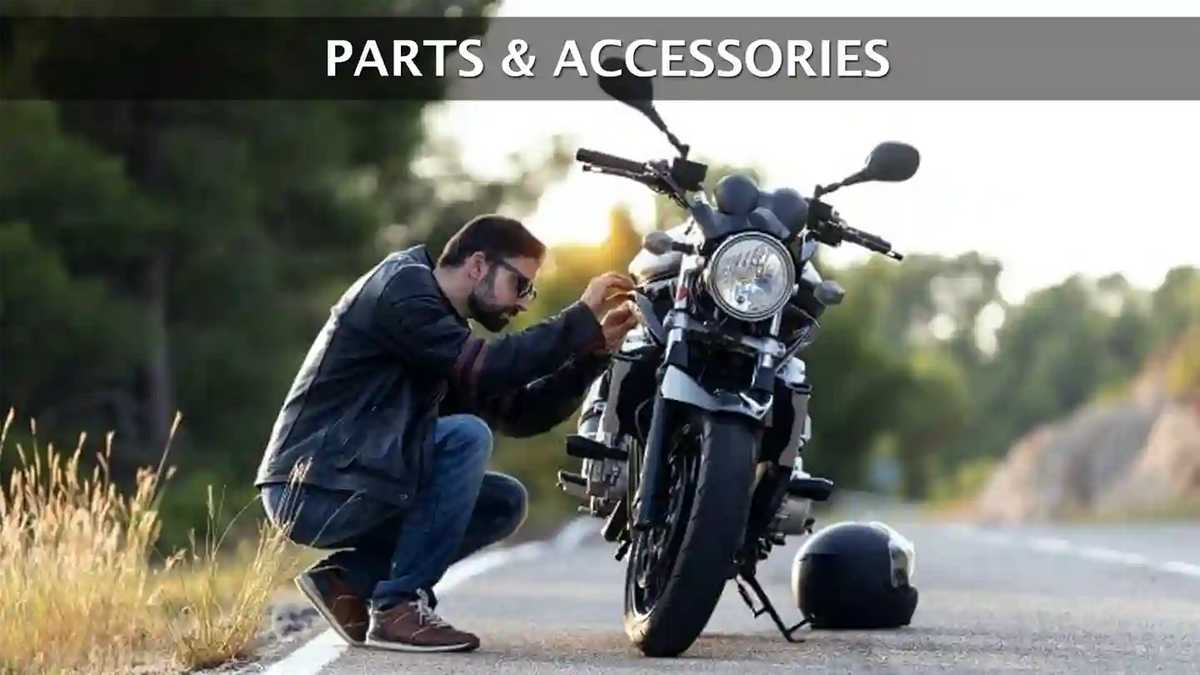 A man kneeling by his motorcycle on the side of the road, adjusting a part on the bike. A helmet rests on the ground nearby, and the text 'Parts & Accessories' is displayed at the top.
