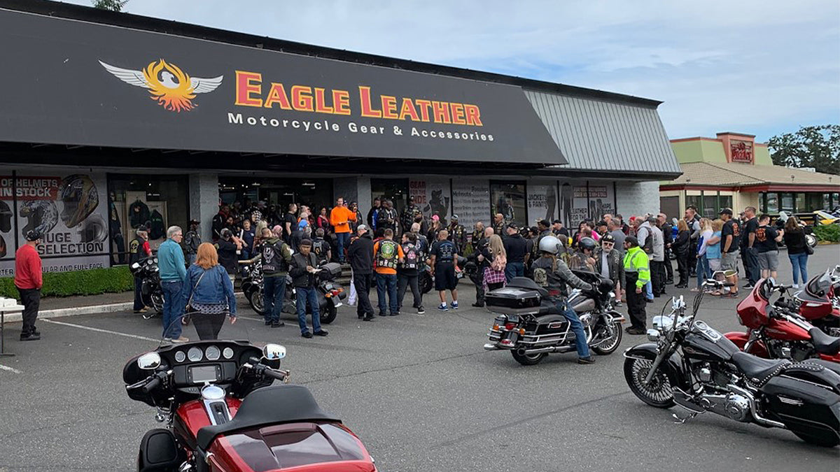 A group of bikers and motorcycles gathered outside the Eagle Leather store, with the logo and 'Motorcycle Gear & Accessories' sign visible above. Riders and customers are seen interacting near the entrance.