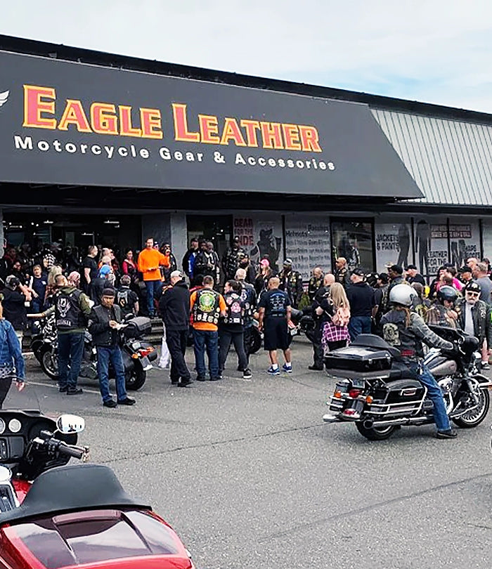 Bikers and motorcycles gathered outside the Eagle Leather store, with the logo and 'Motorcycle Gear & Accessories' sign visible above. The crowd interacts near the entrance, and motorcycles are parked in front.