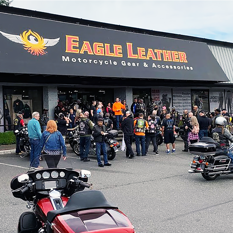 A group of bikers gathered outside the Eagle Leather store, with the logo and 'Motorcycle Gear & Accessories' sign overhead. Motorcycles are parked in front, and people are interacting near the entrance.